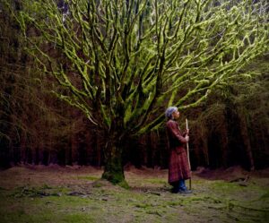 A woman with a staff standing beneath a moss-covered tree